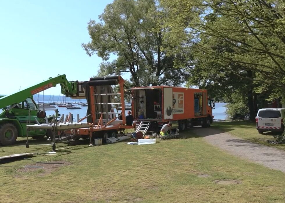 Eine Baustelle am See mit Maschinen und einem Wohnwagen, der aufgestellt wird.