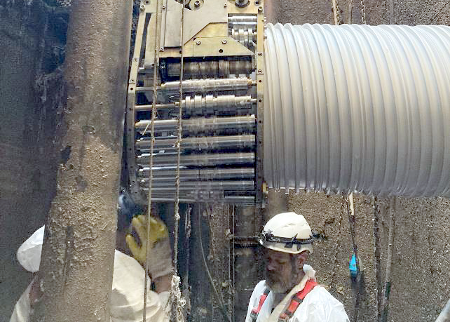 A worker in a hard hat stands beside a drilling machine and large corrugated pipe inside a narrow excavation shaft.