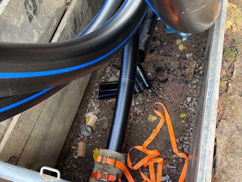 Black utility cables and pipes with blue stripes laid in a trench with orange straps and construction debris.