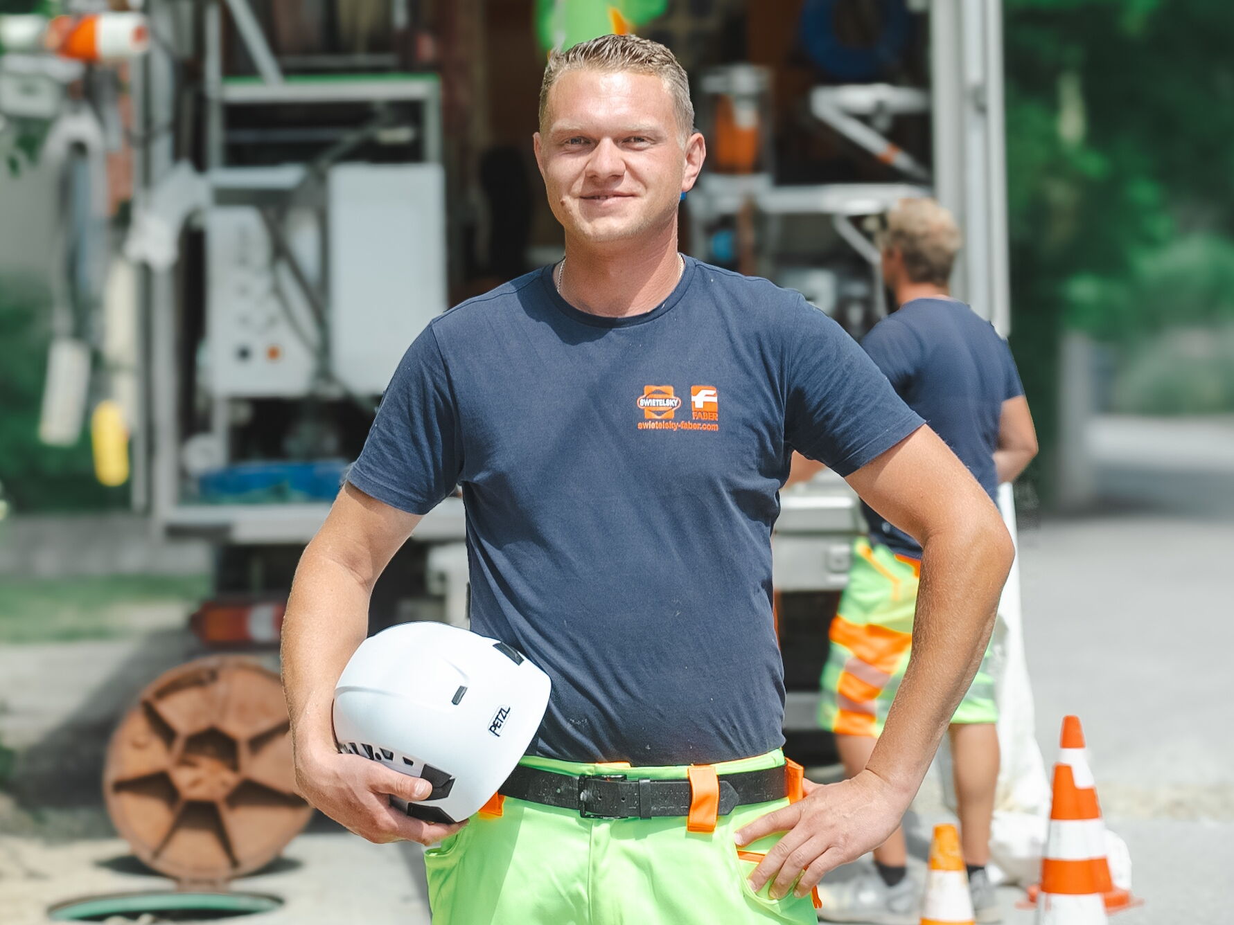 Smiling utility worker holding a white safety helmet, standing on a city street near construction cones and equipment