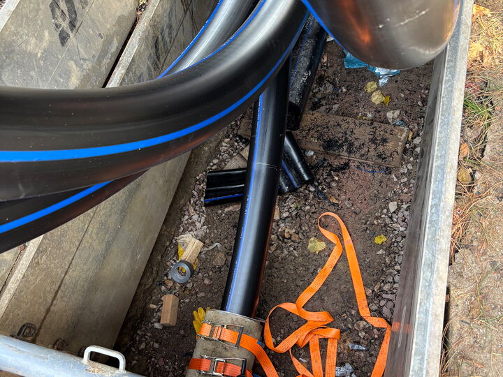 Black utility cables and pipes with blue stripes laid in a trench with orange straps and construction debris.