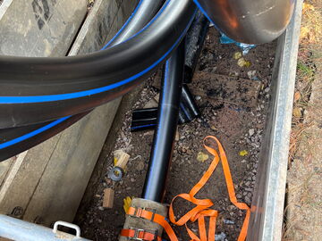 Schwarze Versorgungsleitungen liegen in einem Graben, mit orangefarbenen Gurten und Bauschutt.