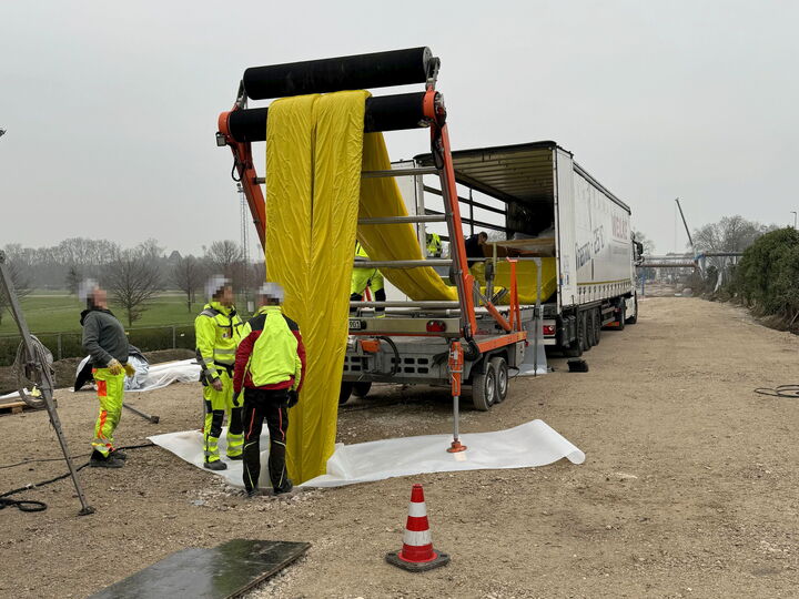 Workers in safety gear unload a large yellow fabric roll from a truck beside a rural road.