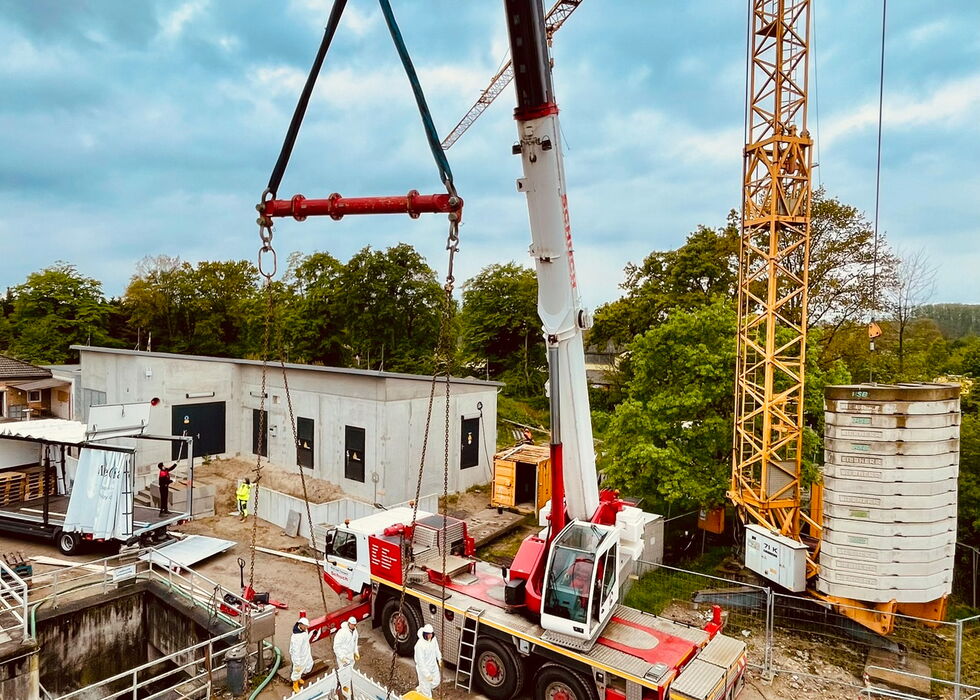 Ein Turmdrehkran hebt schwere Bauteile über ein Betonfundament auf einer Baustelle bei blauem Himmel.