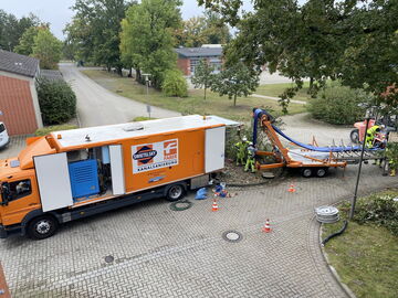 Oranger Versorgungswagen mit Kabelverlegeanhänger und Arbeitern an einer Wohnstraße.