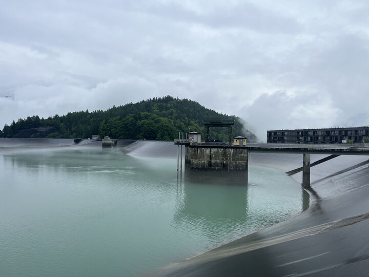 A misty reservoir beside a dam with water spraying from outlets and a forested hill in the background.