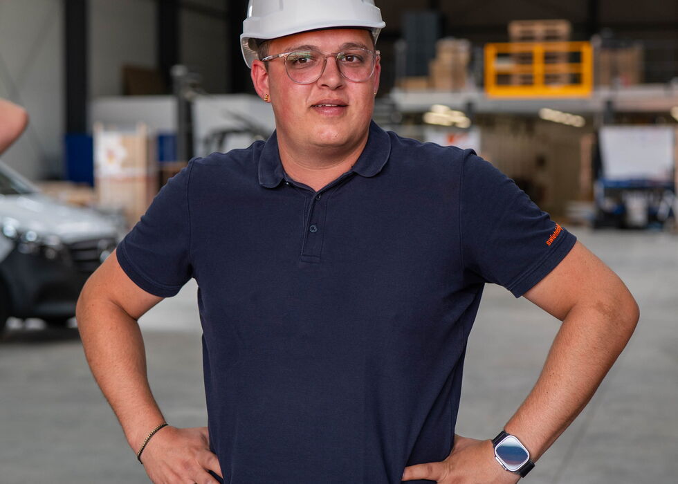 Ein Mann mit Schutzhelm steht mit den Händen in die Hüften gestützt in einer Lagerhalle.