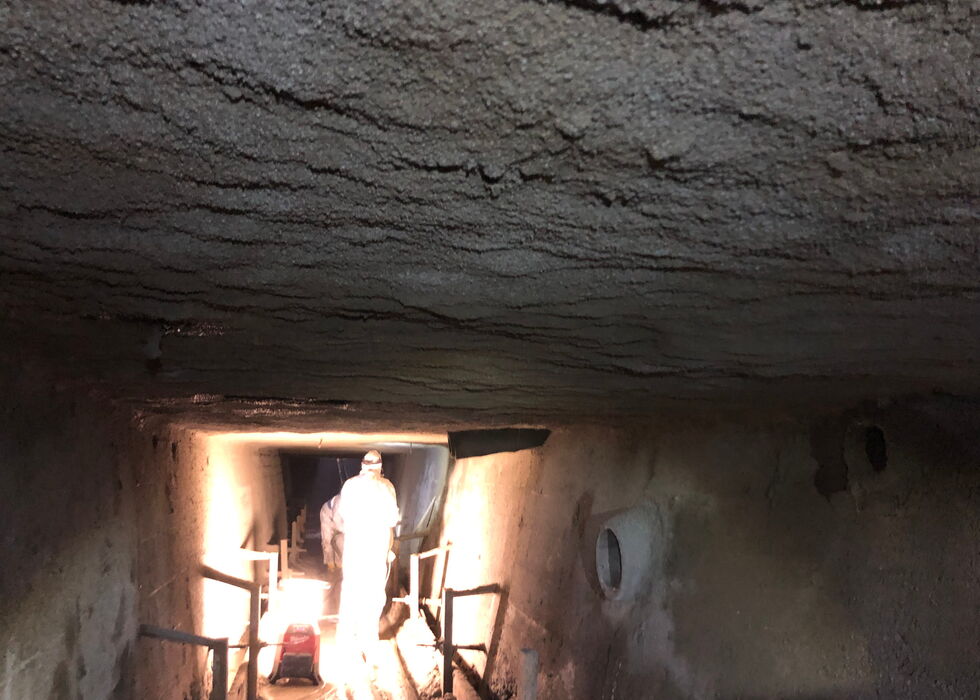 Person wearing protective suit standing in a dimly lit narrow tunnel with concrete walls and ceiling