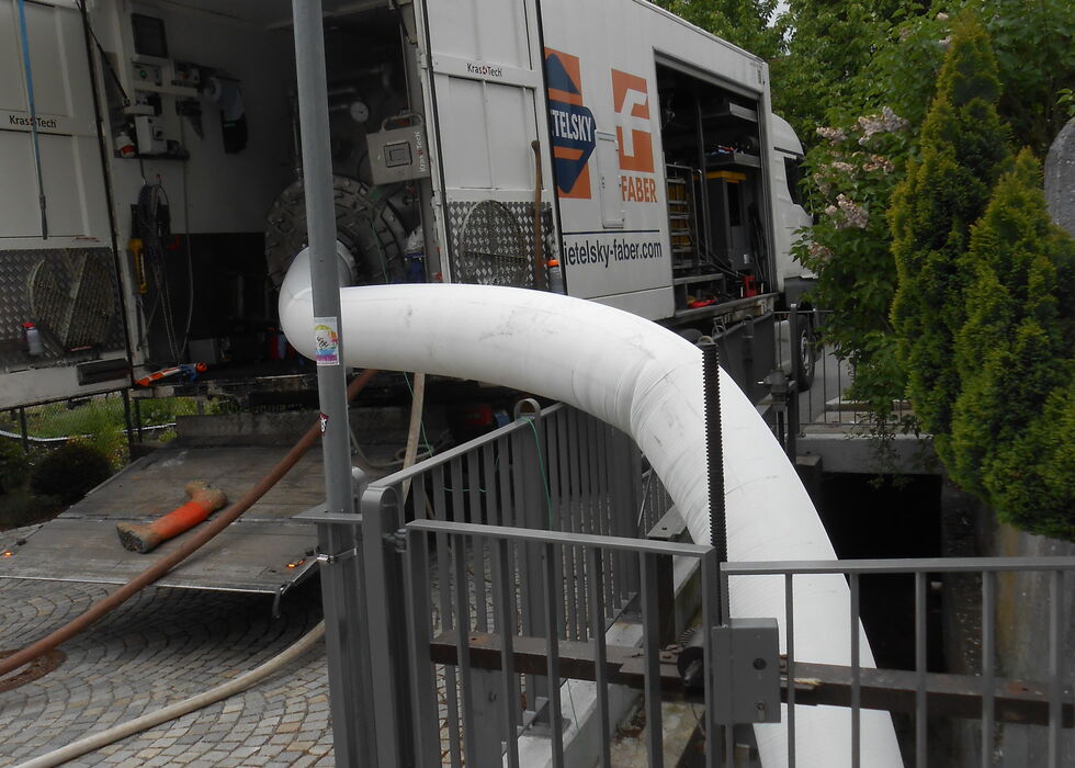 A large white hose from a truck runs into a trench beside a gated sidewalk.
