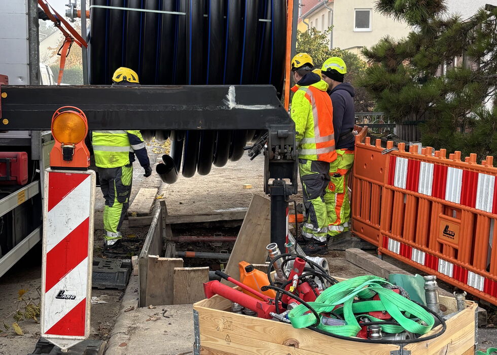 Bauarbeiter mit Warnschutzkleidung helfen dabei, eine große schwarze Kabeltrommel auf einen Tieflader zu heben und zu sichern.