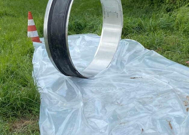 Ein großer Metallring steht aufrecht auf einer Plastikfolie neben einem Grasweg und einem Verkehrskegel.