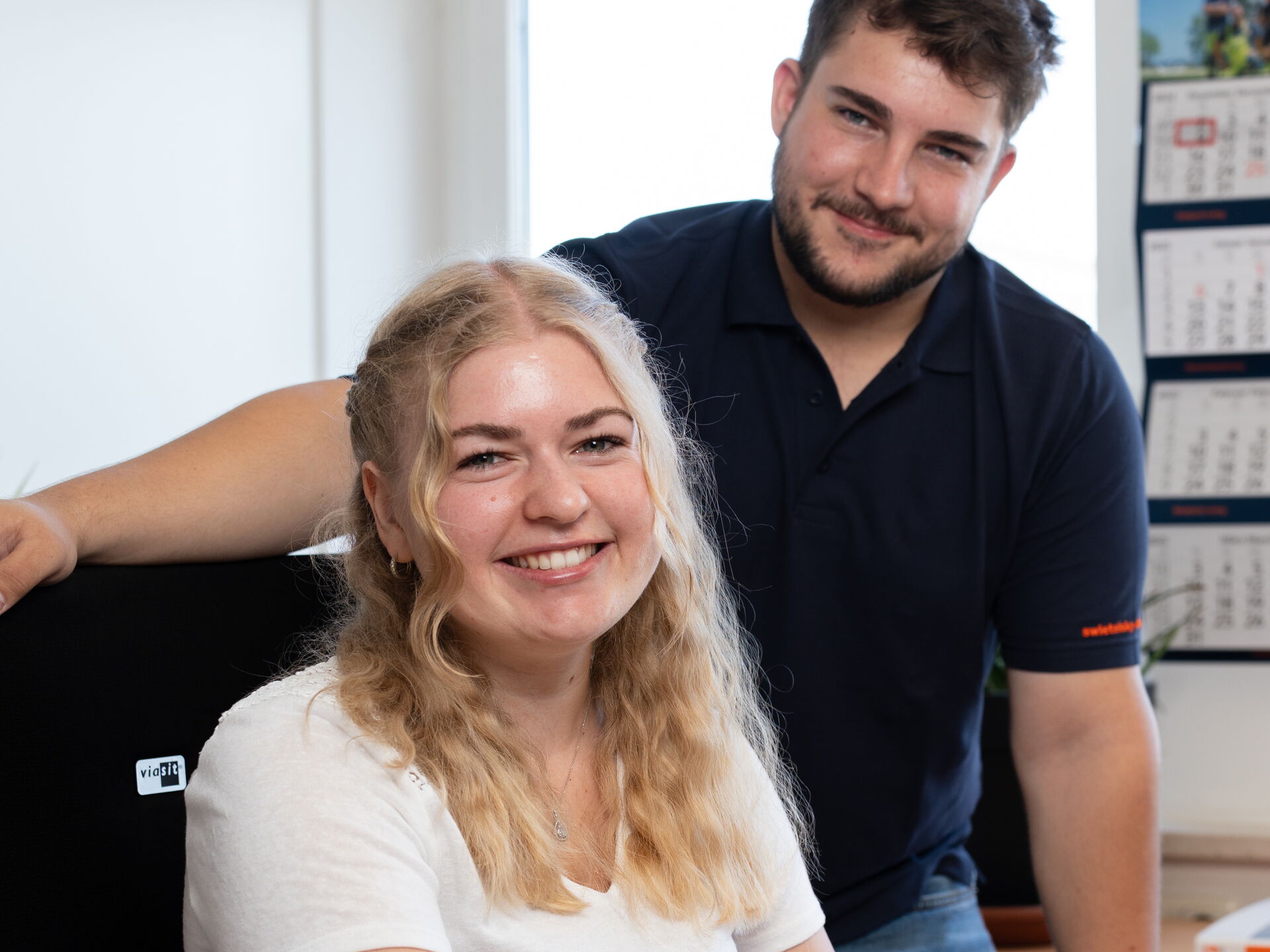 Lächende junge Frau sitzt am Schreibtisch und benutzt Computermaus, ein Mann steht hinter ihr in einer lockeren Büroumgebung