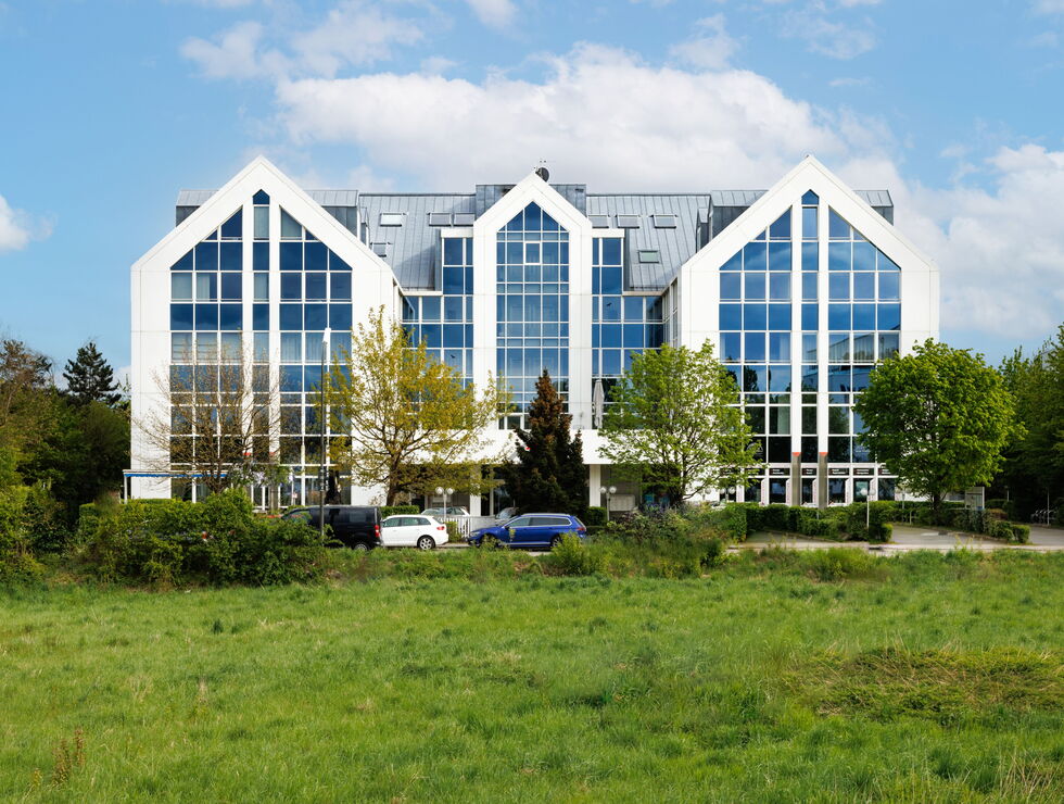 A modern office building with large glass windows and white gabled frames beside a grassy field and trees.
