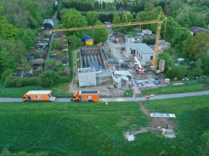 Aerial view of a construction site with a crane, buildings under construction, and trucks beside a road surrounded by fields and trees.