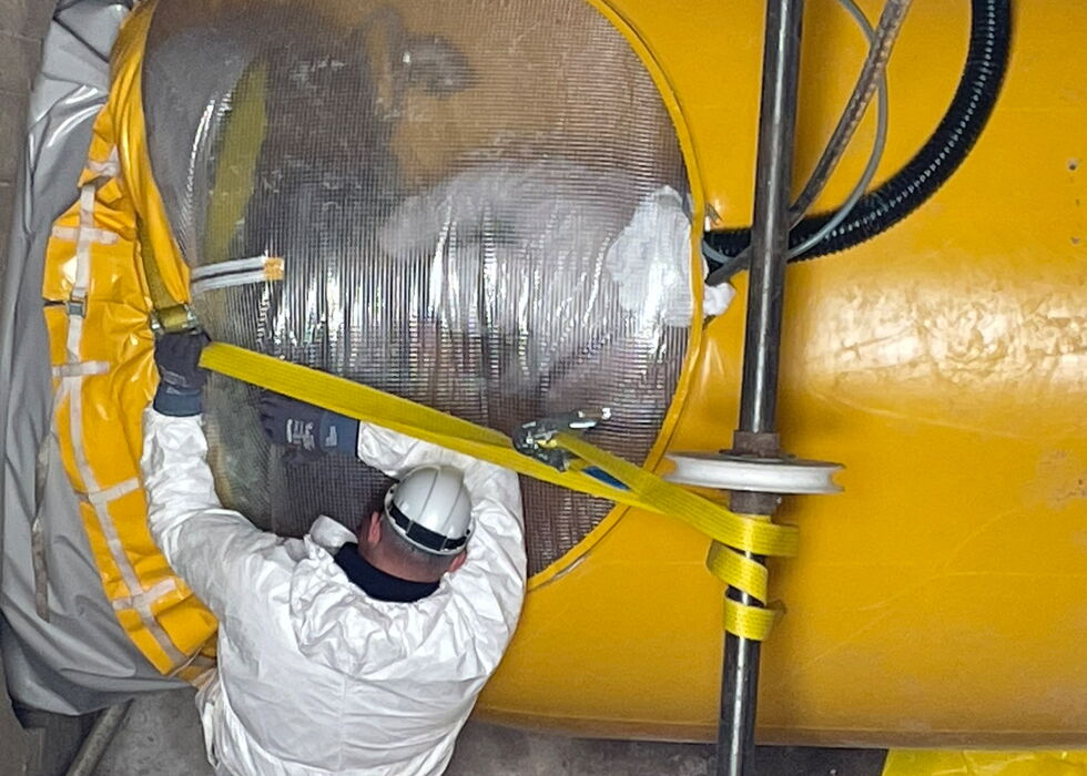Ein Arbeiter in Schutzkleidung inspiziert in einem engen Industriebereich ein großes gelb isoliertes Rohr.
