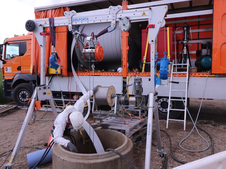 A worker operates a truck-mounted lifting frame beside an orange service truck at a well site.