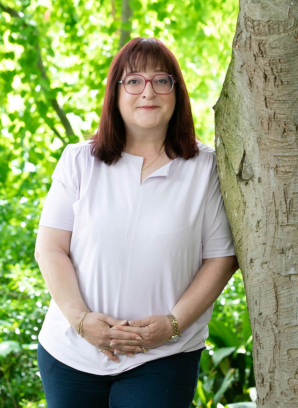 Eine Frau mit Brille steht neben einem Baum in einem grünen Park.