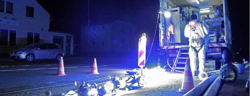 A worker operates equipment at night near a brightly lit construction zone with cones and machinery.