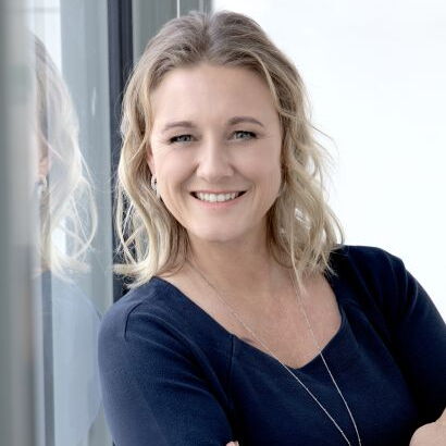 Eine fröhliche Frau mit blonden Haaren steht neben einem Fenster und lächelt selbstbewusst mit verschränkten Armen.