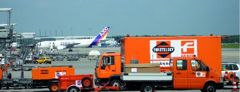 Eine belebte Flughafenszene mit orangefarbenen Servi fahrzeugen und einem Passagierflugzeug im Hintergrund.