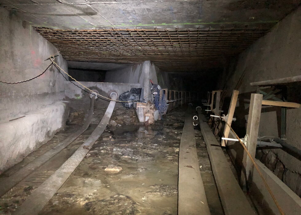 Dimly lit underground tunnel with water on the floor, pipes on the right, and construction materials scattered