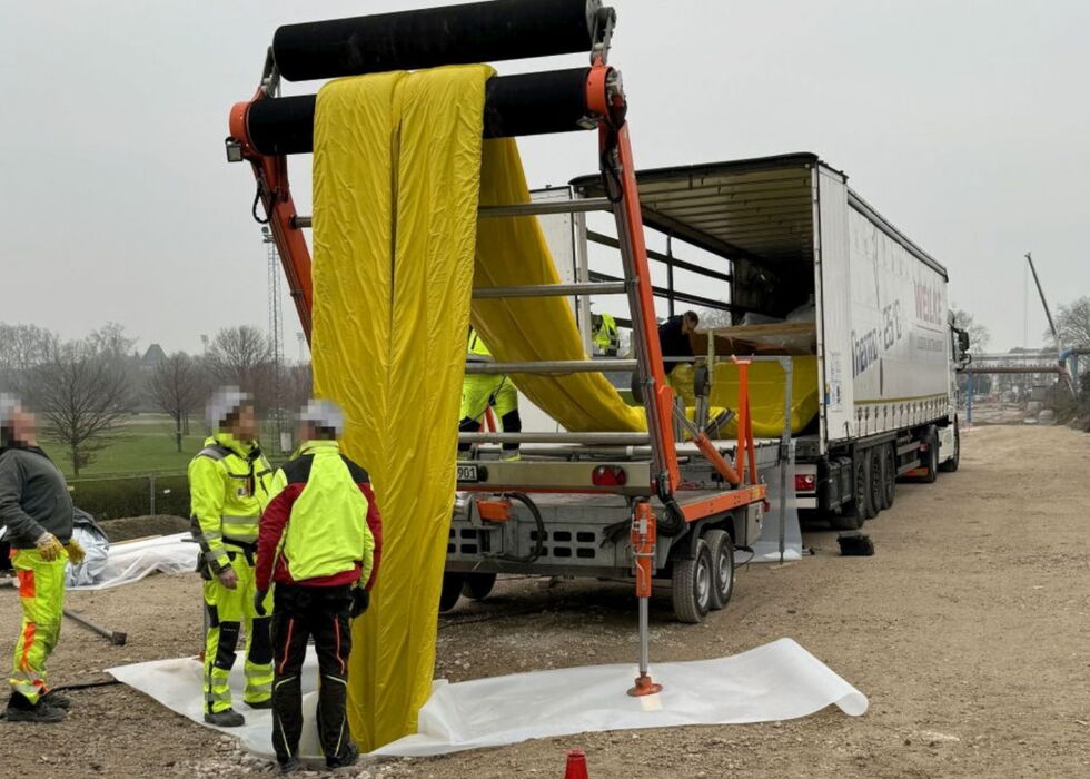 Arbeiter verwenden ein Lkw-montiertes System, um gelbe Schutzplanen an einer Baustelle zu laden.
