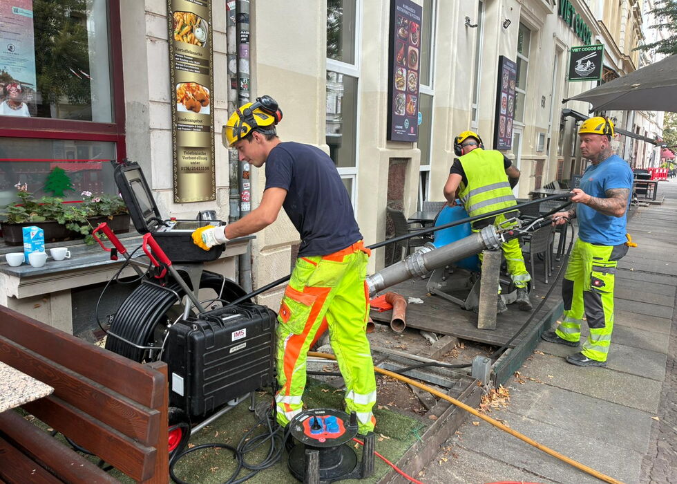Arbeiter in Warnkleidung nutzen Schläuche und Geräte für Kanalarbeiten auf einem städtischen Gehweg.