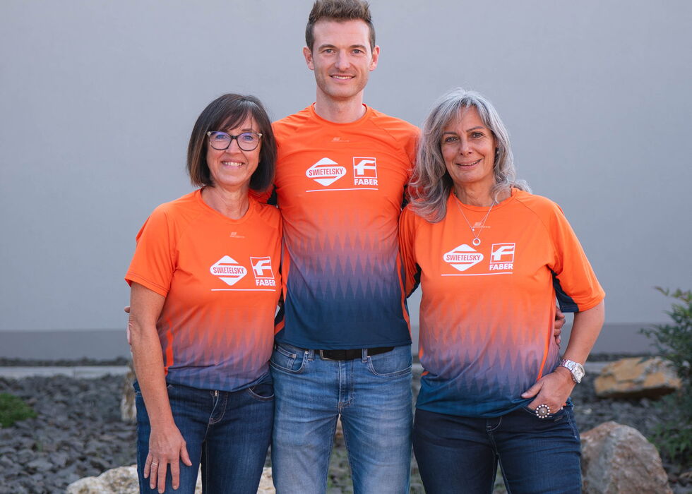 Drei lächelnde Personen stehen draußen zusammen und tragen passende orangefarbene Teamshirts und Jeans.