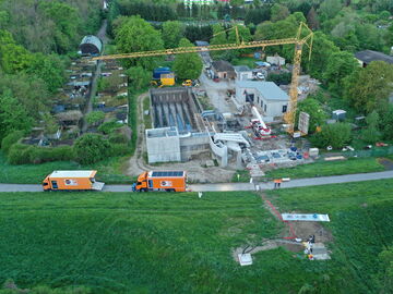 Luftaufnahme einer Baustelle mit Kran, im Bau befindlichen Gebäuden und Lastwagen neben einer Straße, umgeben von Feldern und Bäumen.