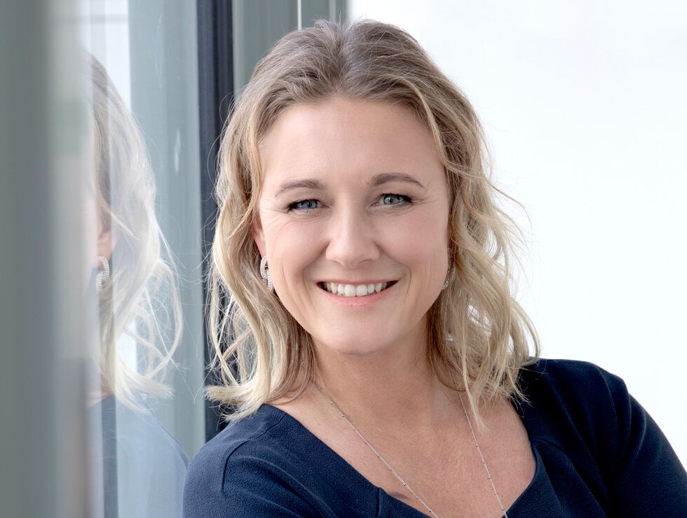 Portrait of a smiling blonde woman standing by a window with her arms crossed.