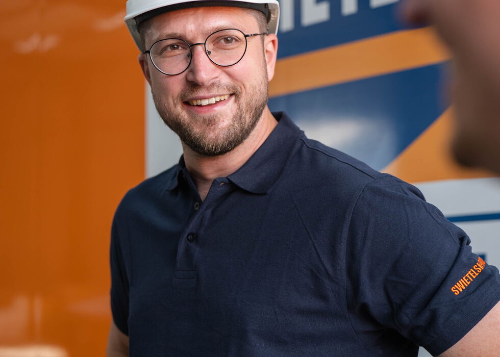 Lächelnder Mann mit weißem Schutzhelm und dunkelblauem Polohemd vor einem orangefarbenen LKW.