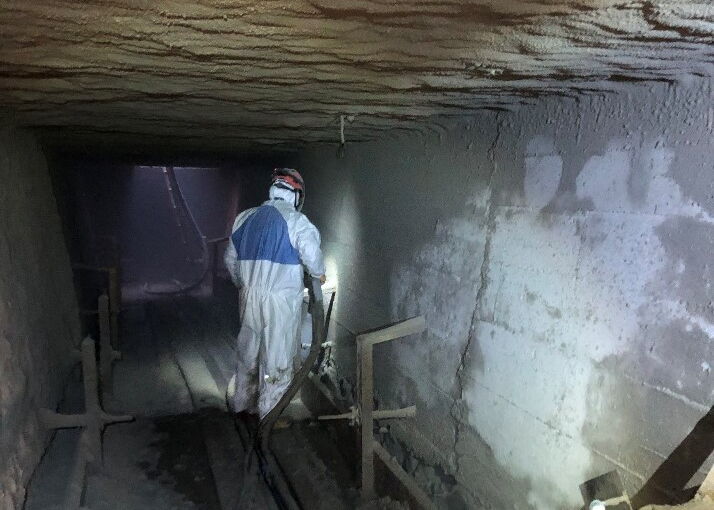 Person in protective clothing walking through a dimly lit underground mine tunnel with industrial equipment
