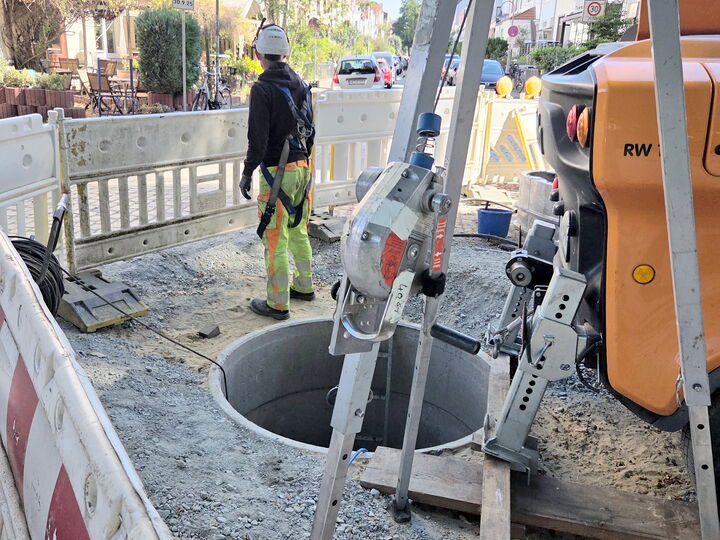 Construction worker at a street dig site beside an open manhole and drilling rig on a city road.