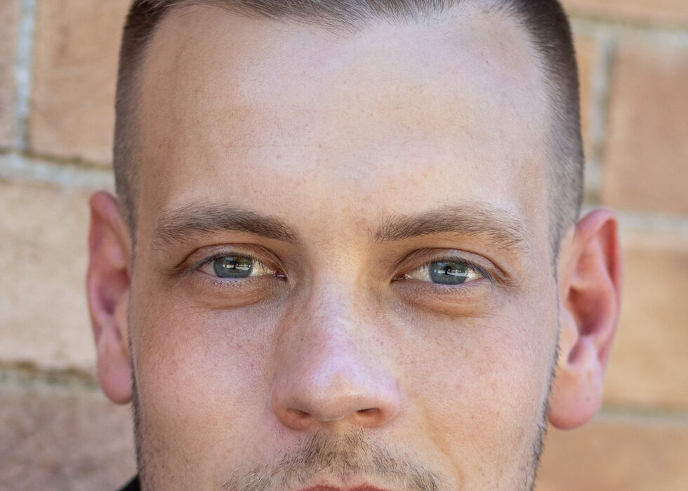 Close-up of a young man with blue eyes and slicked-back hair standing in front of a brick wall