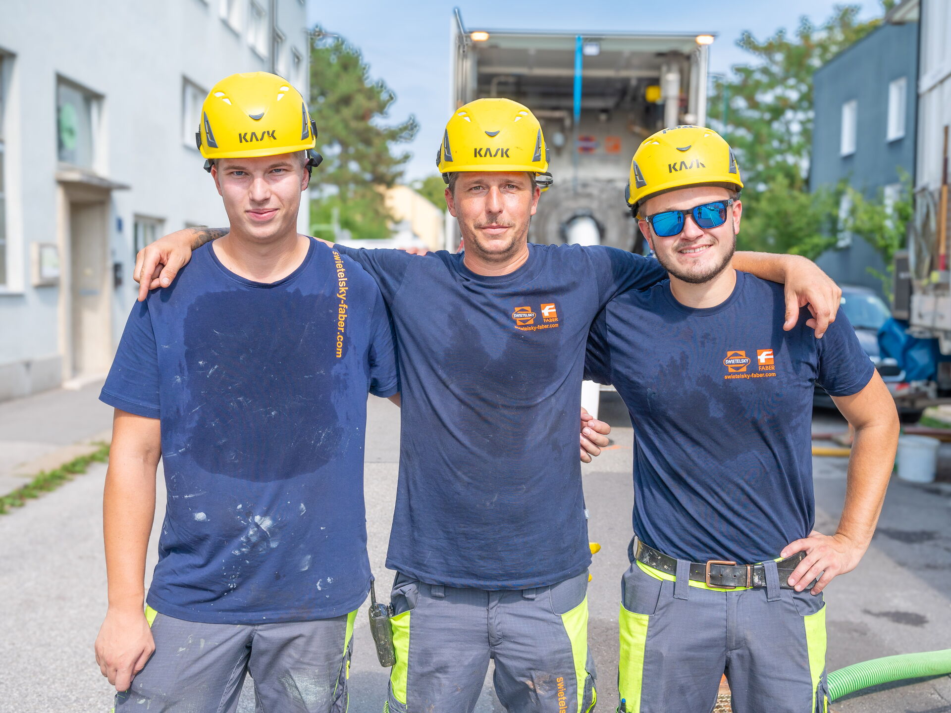 Three construction workers wearing yellow helmets and navy blue shirts standing with arms around each other on a street with trucks in the background