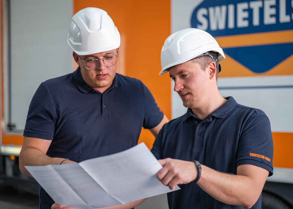 Zwei Bauarbeiter mit Schutzhelmen prüfen gemeinsam einen Bauplan auf der Baustelle.