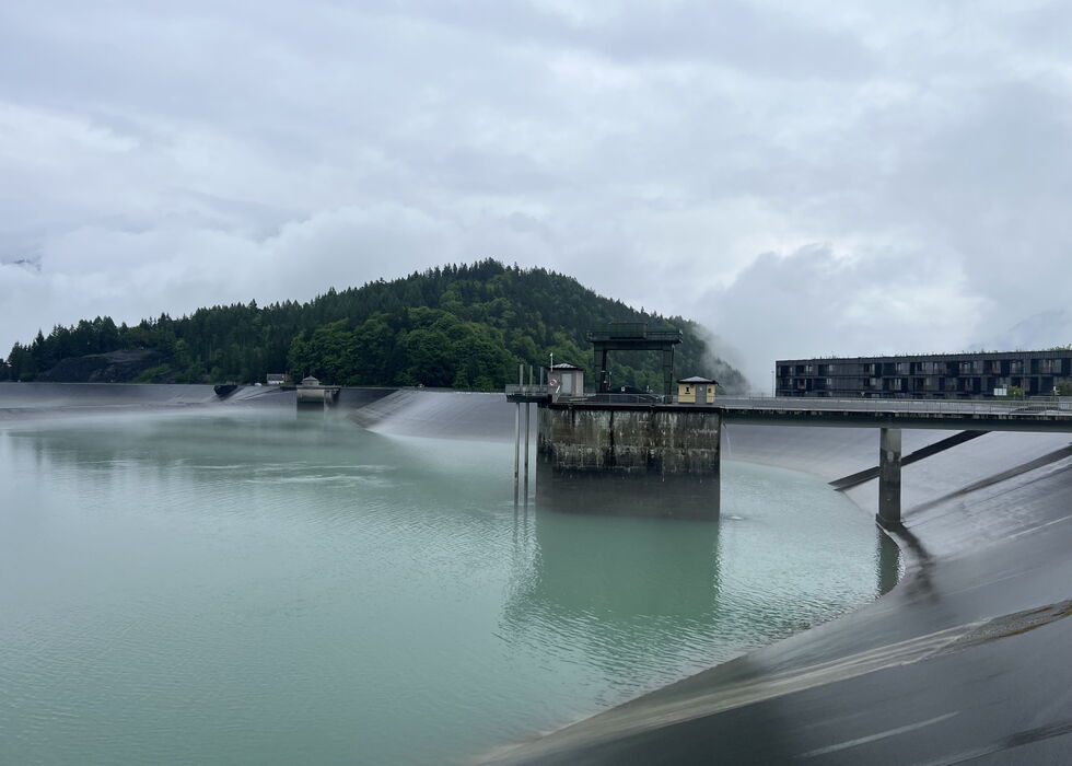 A misty reservoir beside a dam with water spraying from outlets and a forested hill in the background.