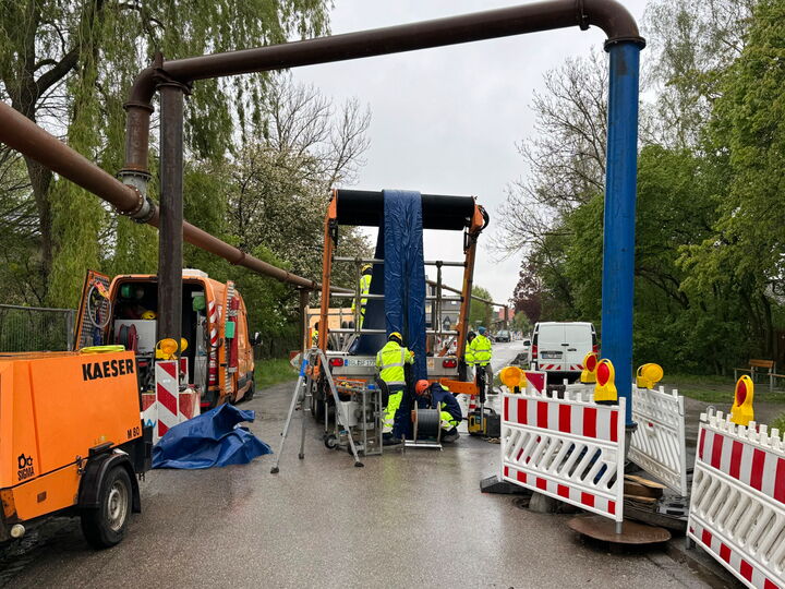 Workers and barriers around machinery repairing a pipeline crossing on a wet road.