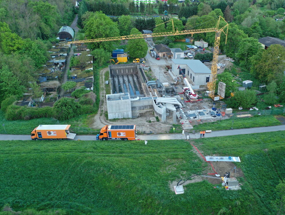 Aerial view of a construction site with a crane, buildings under construction, and trucks beside a road surrounded by fields and trees.