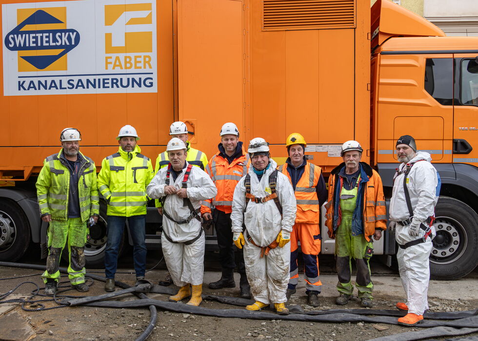 Mehrere Arbeiter in Schutzkleidung stehen vor einem orangefarbenen Kanalreinigungsfahrzeug.
