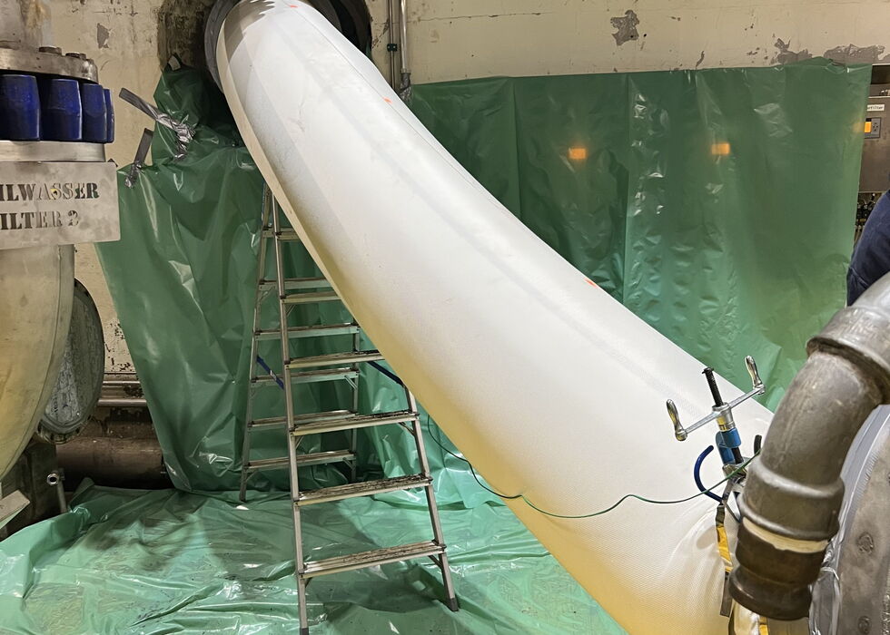 White flexible duct installed through a wall opening in an industrial room, with a ladder and green sheeting nearby.