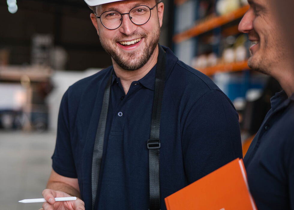 Zwei Lagermitarbeiter mit Schutzhelmen sprechen und prüfen gemeinsam ein Tablet und einen Ordner im Lager.