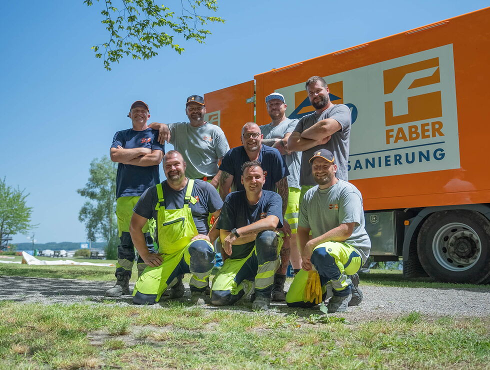Group of workers posing in front of an orange delivery/utility truck outdoors on a sunny day.