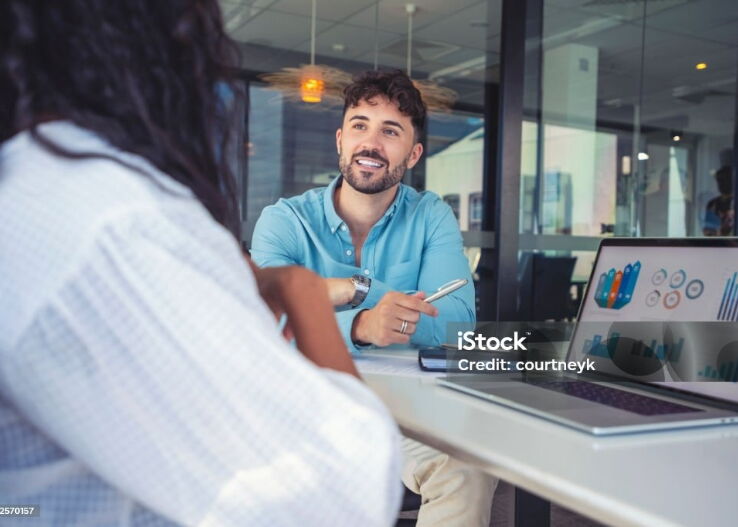 Ein junger Mann lächelt, während er mit einer professionellen Frau spricht, die mit einem Laptop und Diagrammen in einem modernen Büro sitzt.
